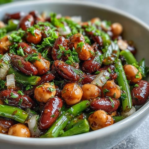 A close-up of a vibrant Three-Bean Salad in a white bowl, featuring green beans, kidney beans, and chickpeas glistening with tangy vinaigrette.  